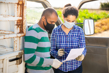 Wall Mural - Hispanic female farmer in medical mask discussing contract with driver of transport company near car on farm field. Pandemic prevention and social distancing concept