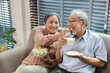 © feeling lucky - Happy smiling asian senior man and woman sitting on sofa and eating popcorn while having fun with movie rest indoor at home living room. Couple elder husband and wife embrace are happy while watch TV