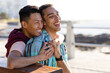© Wavebreak Media - Happy biracial gay male couple sitting on bench and embracing on promenade by the sea