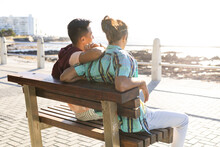 Happy Biracial Gay Male Couple Sitting On Bench And Embracing On Promenade By The Sea