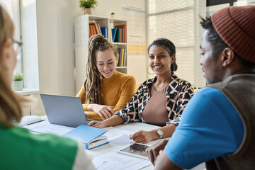 Smiling girl talking to teacher while sitting at table with her classmates during lesson in class