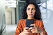 © Liubomir - Upset thinking business woman close up inside office, young hispanic woman reading bad news on phone online, business woman at workplace holding smartphone typing text message.