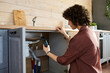 © pressmaster - Side view of young housewife taking photo of broken pipe on smartphone while squatting in front of open kitchen counter at home