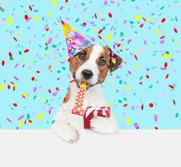 Canvas Print - Jack russell terrier puppy wearing a party cap blows into party horn and holds gift box above empty white banner