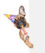 French Bulldog puppy wearing party cap blows into party whistle and looks from behind empty white banner. isolated on white background