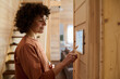 © pressmaster - Young pretty woman with dark curly hair pointing at tablet screen hanging on wooden wall while choosing settings of alarm system of house