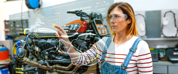 Concentrated female mechanic using hud panel screen with augmented reality hologram to review motorcycle on factory. Futuristic user interface concept.