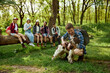 © master1305 - Young man playing with dog. Group of young people, friends resting in forest, going hiking on warm sunny day, sitting on big fallen tree. Active lifestyle, nature, sport and hobby, friendship concept
