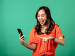 © Daniel - A portrait of an Asian woman wearing an orange shirt, looking surprised while holding a smartphone, isolated on a green background.