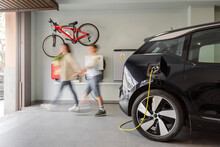 Electric Vehicle Charging Station In A Private Home With An Unrecognizable Blurred Family Leaving The House, With A Bicycle Hanging On The Wall. Copy-space, Horizontal., Motion Blur.