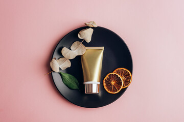 Wall Mural - Golden cream tube and dry orange slices, eucalyptus leaves on black plate on a pink background. Top view, flat lay, mockup