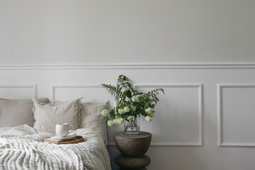 Wall Mural - Breakfast in bed. Cup of coffee, wicker tray. Green bouquet of white viburnum, fern and solomons seal flowers. Modern boho night stand. Bedroom view. Beige pillows, linen blanket. Elegant moulding.