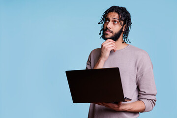 Arab man reading programming report on laptop while looking away with puzzled facial expression. Person rubbing chin while thinking about cyber security and holding portable computer