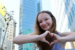 beautiful girl shows heart with hands against backdrop of skyscrapers and big city with huge glass buildings straighten hair relax smile travel like 