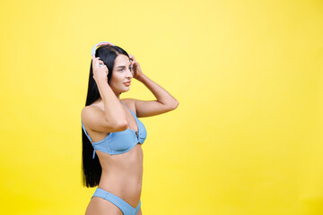Happy young woman wearing in blue swimsuit listening music in headphones and dancing, singing song and having fun isolated on yellow wall background studio. Summer rest concept