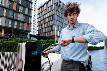 Businessman with smartwatch at modern charging station for electric vehicle with background of residential buildings as concept for progressive lifestyle of using eco-friendly as alternative energy.