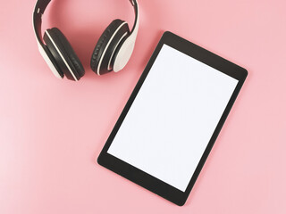 flat lay of tablet with white blank screen and headphones on pink background.