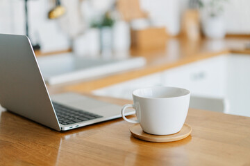 Cup of cofee and laptop on wooden table, view on white kitchen in modern style