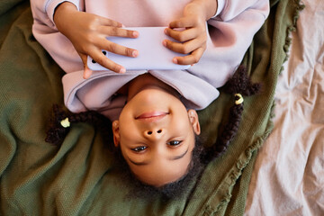 Wall Mural - Top view portrait of black girl looking at camera upside down while lying on bed with smartphone