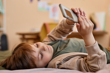 Wall Mural - Portrait of cute boy holding smartphone while lying on bed and smiling, gen Alpha