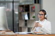 © NAMPIX - Young confident businesswoman with headphone holding coffee cup smiling and looking at desktop monitor screen while sitting at desk office.