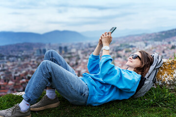 Young adult happy woman traveler lying on green grass on top overlooking european city using smartphone