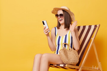 Satisfied positive woman wearing striped swimwear and hat sitting on deck ?hair resting at resort having video call waving hand saying hello.