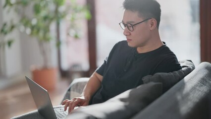 Pensive asian man in eyeglasses typing on laptop at home