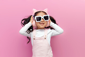 little asian girl in pink children's headphones and glasses listens to music and dances on pink isolated background