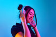 Portrait of beautiful young korean girl listening to music in headphones against blue studio background in neon light. Concept of emotions, facial expression, youth, lifestyle, inspiration, sales, ad
