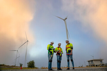 Two male engineers and female foreman team talk to blueprints, planning work to inspect and repair wind turbines to produce electricity in the middle of the turbine field.