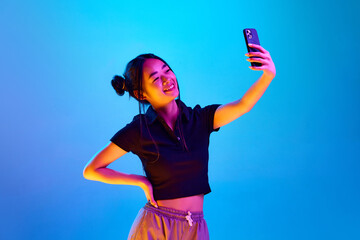 Wall Mural - Portrait of smiling, young, korean girl taking selfie with mobile phone against blue studio background in neon light. Concept of emotions, facial expression, youth, lifestyle, inspiration, sales, ad