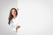 © Prostock-studio - Smiling senior woman medical worker looking out behing white advertising board with free space for text or design