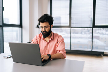 Portrait of Indian freelancer male in casual clothes working on laptop sitting at table in light coworking office on background of window. Bearded business man in glasses looking at computer screen.
