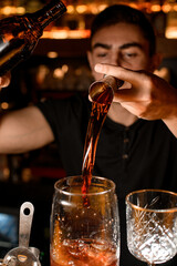 Wall Mural - Bartender masterfully pours strong alcohol from steel jigger into transparent mixing cup with ice cubes