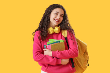 Wall Mural - Female student with headphones, notebooks and backpack on yellow background