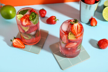 Glasses of fresh strawberry mojito with shaker on blue table near orange wall