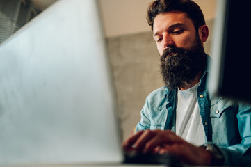 A data analyst is sitting at the home office and typing on a laptop surrounded by monitors.