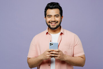 Wall Mural - Young smiling happy Indian man he wear pink shirt white t-shirt casual clothes hold in hand use mobile cell phone look camera isolated on plain pastel light purple background studio Lifestyle concept