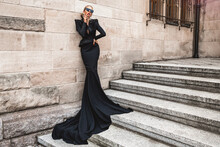 Elegant Luxury Evening Fashion. Glamour, Stylish Elegant Woman In Black Long Evening Gown Dress Is Posing In The City Outdoor. Female Model In Amazing Long Dress. Outdoor Shoot. Vogue. Couture.