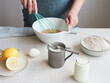 Woman beating eggs with a whisk in a bowl. Ingredients to prepare a dessert or pastry recipe. Flour, milk and lemon on the table.