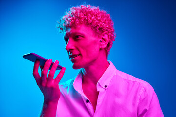 Portrait of man with curly hair recording voice message with phone against blue studio background in pink neon light. Concept of human emotions, lifestyle, youth, facial expression, communication