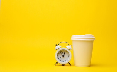 White disposable cardboard cup of coffee with alarm clock on yellow background. Mockup