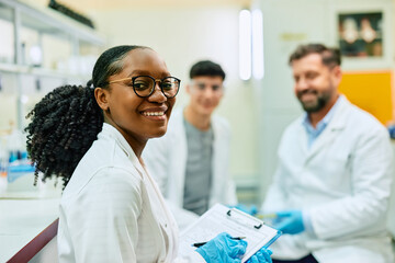 Wall Mural - happy black biochemist working with her colleagues in lab and looking at camera.