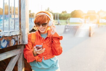 Wall Mural - Smiling stylish woman in bright clothes, sunglasses wearing wireless headphones on her head and seting phone for listening music. Walk in city skate park on sunset. Hipster lifestyle. Selective focus.