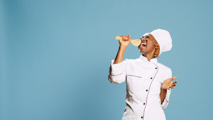 Wall Mural - Young happy chef using wooden spoon to sing in studio, having fun singing with cooking utensils on camera. Professional cook with apron playing aroung and holding kitchen tool.