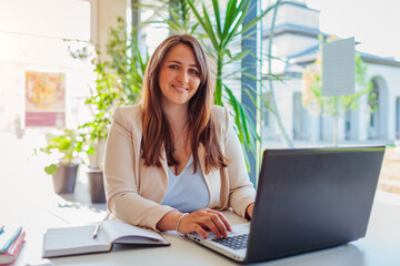 Happy businesswoman working online in office using laptop. Smiling middle-aged woman looking at camera sitting at desk