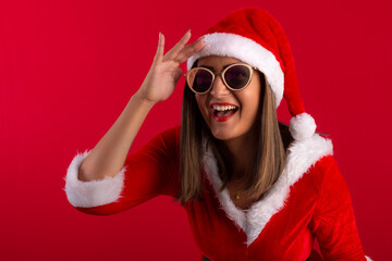 Very pretty adult woman wearing sunglasses, wearing Santa's hat and dress, in studio shot with red background, with space for text and making various facial expressions