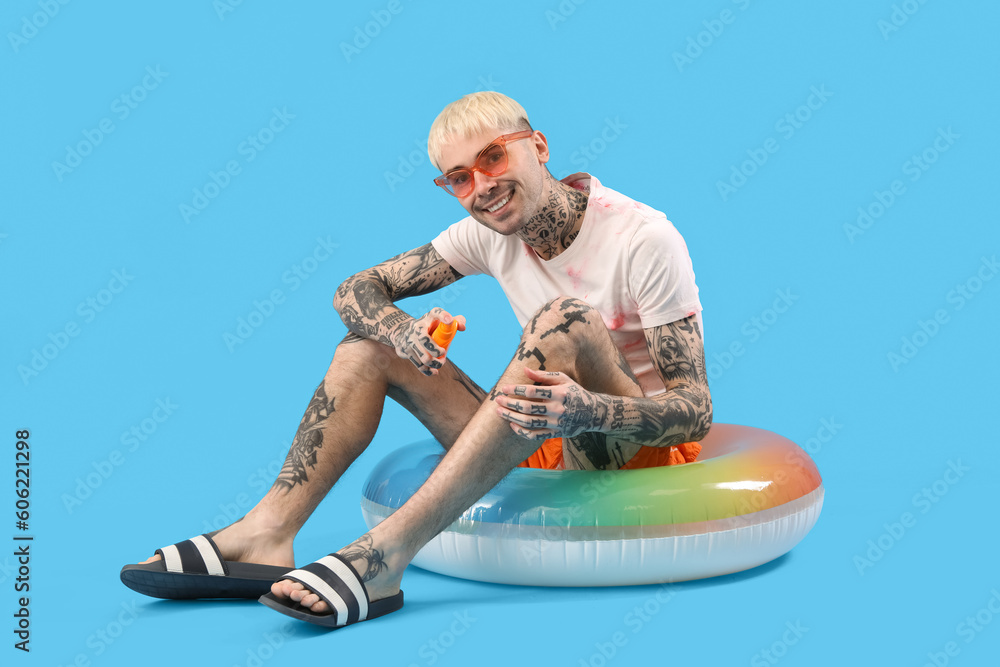 Tattooed man with sunscreen cream and swimming ring on light blue background