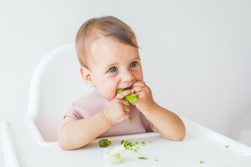 baby little girl 8 months old sits in a high chair and eats complementary foods green broccoli, close-up portrait looks at the camera. baby food concept